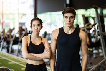 Fototapeta na wymiar Portrait of young handsome fit Caucasian man and beauty Asian woman standing showing muscle body in gym or fitness club with exercise equipment in background. Working out for health, well being.
