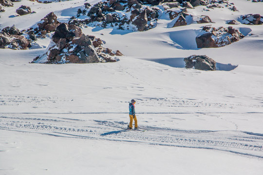 Snowboarder going down the slope of mount Elbrus. He wore bright clothes: yellow pants and a turquoise jacket, with a helmet on his head. The picture was taken in November.
