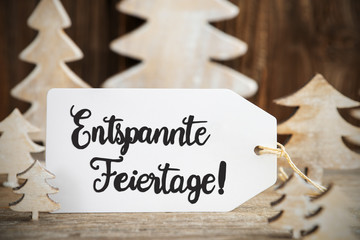 Label With German Text Entspannte Feiertage Means Merry Christmas. White Wooden Christmas Tree As Decoration. Brown Wooden Background