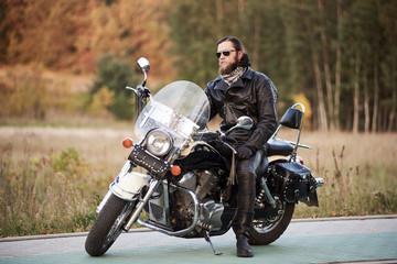Young bearded biker in black leather clothing and sunglasses sitting on cruiser motorcycle on clean paved roadside on background of vintage trees golden bokeh foliage.