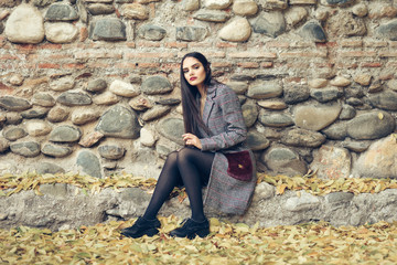 Beautiful girl wearing winter coat sitting on the floor of an urban park full of autumn leaves.