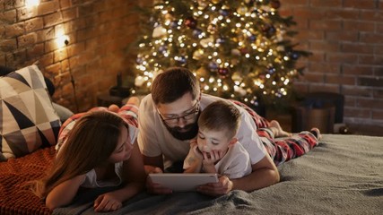 Happy father with little son and daughter in pajamas lying in bed at home, uses digital tablet computer before sleeping. Christmas tree and lights in the background - Powered by Adobe