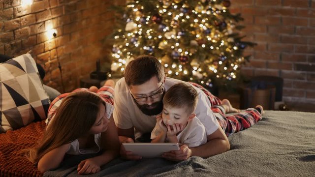 Happy father with little son and daughter in pajamas lying in bed at home, uses digital tablet computer before sleeping. Christmas tree and lights in the background
