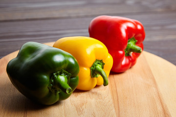 Red, green, and yellow bell peppers, cutting board on wooden background. Three sweet peppers in different colors on brown table, vegetable ingredient, healthy food