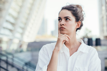 Portrait of calm and thoughtful young woman posing on camera. Stand and look to left. Modern buildings behind. Stand alone outside. Pretty beautiful woman.