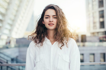 Pretty calm young woman posing straight on camera. Stand alone outside. White buildings behind. Wear white stylish blouse. Daylight.