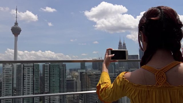 4K, Asian woman take photo with smartphone of famous Kuala Lumpur communications tower Menara KL. Tourists visit and picture an antenna. Television tower skyline in Malaysia-Dan