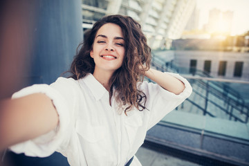 Positive nice young woman with dark curly hair in white blouse posing while taking selfie. Smiling. Alone outside. Beautiful buildings. Sunset.