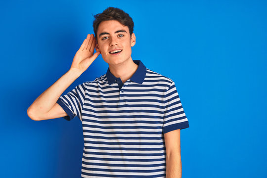 Teenager boy wearing casual t-shirt standing over blue isolated background smiling with hand over ear listening an hearing to rumor or gossip. Deafness concept.