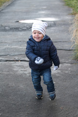 Funny toddler boy running on a walk in autumn. Smiling child wearing white hat, blue jacket, woolen mittens and jeans on sidewalk. Happy kid portrait