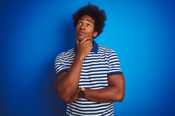 Fototapeta na wymiar African american man with afro hair wearing striped polo standing over isolated blue background with hand on chin thinking about question, pensive expression. Smiling with thoughtful face. 
