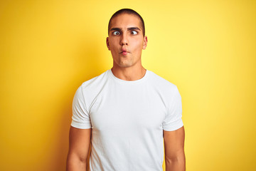 Young caucasian man wearing casual white t-shirt over yellow isolated background making fish face with lips, crazy and comical gesture. Funny expression.