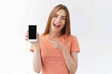Fototapeta premium Pretty, cheerful young caucasian woman in striped t-shirt holding smartphone, pointing black mobile phone display and smiling as discuss new app, recommend follow her social media, white background