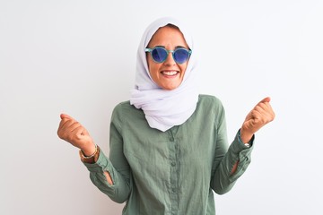 Young Arab woman wearing hijab and summer sunglasses over isolated background celebrating surprised and amazed for success with arms raised and open eyes. Winner concept.