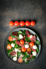 Salad with prosciutto, mozzarella, cherry tomatoes, capers and spinach on a dark plate on a dark background top view copy space.