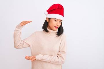 Obraz na płótnie Canvas Young beautiful chinese woman wearing Christmas Santa hat over isolated white background gesturing with hands showing big and large size sign, measure symbol. Smiling looking at the camera. Measuring 