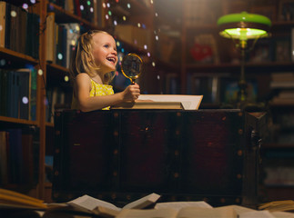 A little girl at home library reading a book sitting in an old chest holding a magnifying glass. Children and education many books on the floor, night time