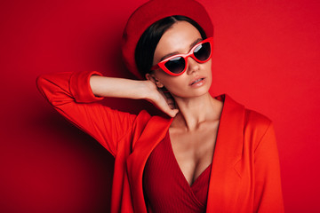 Portrait of young brunette woman isolated over red background. Stand alone and posing. Hold hand on neck. Wear red jacket, beret and sunglasses. Slim stylish model.