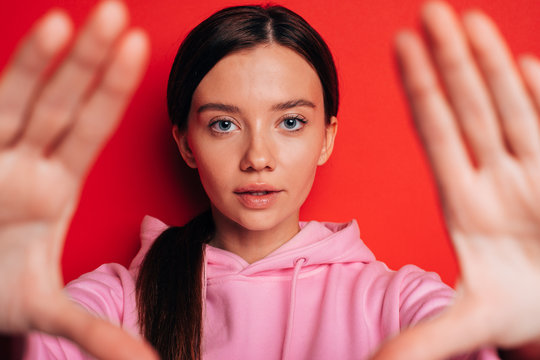 Calm peaceful young woman look on camera straight. Show hands close to shot. Wear pink hoody. Brunette isolated over red background.