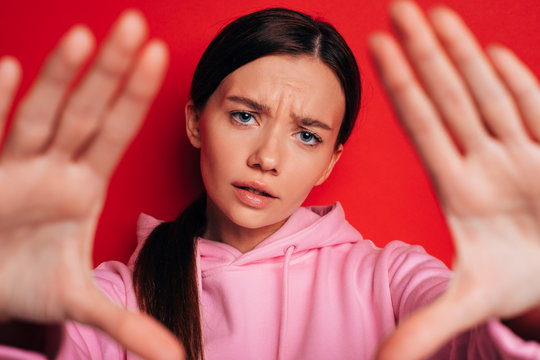Worried young woman in pink hoody look straight. Show palms of hands. Posing in modern way. Disturbed. Isolated over red background.
