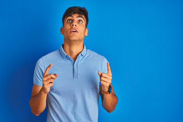 Young indian man wearing casual polo standing over isolated blue background amazed and surprised looking up and pointing with fingers and raised arms.