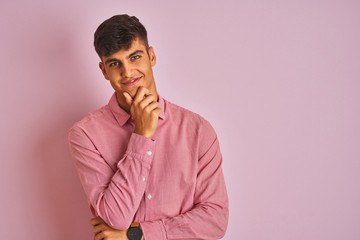 Young indian man wearing elegant shirt standing over isolated pink background looking confident at the camera smiling with crossed arms and hand raised on chin. Thinking positive.