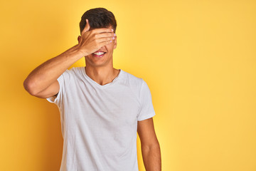 Young indian man wearing white t-shirt standing over isolated yellow background smiling and laughing with hand on face covering eyes for surprise. Blind concept.