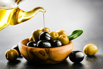 Pour virgin olive oil on green and black original olives in bowl on black background.