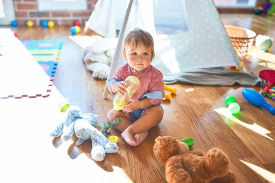 Adorable toddler holding feeding bottle around lots of toys at kindergarten