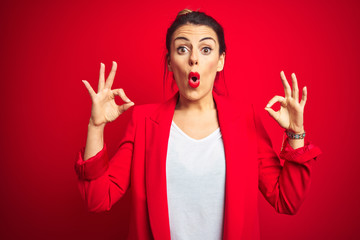 Young beautiful business woman standing over red isolated background looking surprised and shocked doing ok approval symbol with fingers. Crazy expression