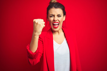Young beautiful business woman standing over red isolated background angry and mad raising fist...