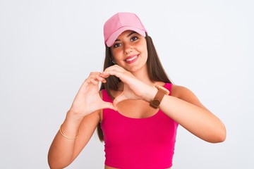 Young beautiful girl wearing pink casual t-shirt and cap over isolated white background smiling in love showing heart symbol and shape with hands. Romantic concept.