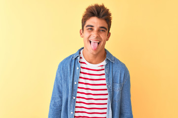 Young handsome man wearing striped t-shirt and denim shirt over isolated yellow background sticking tongue out happy with funny expression. Emotion concept.