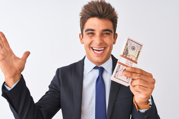 Young handsome businessman wearing suit holding dollars over isolated white background very happy and excited, winner expression celebrating victory screaming with big smile and raised hands