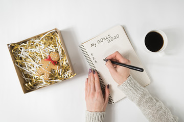 Female hands writing My Goals 2020 in a notebook. Mug of coffee and gingerbread man, top view