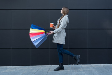 Happy young woman with colorful bags and paper cup walking on the street. Black wall on background