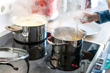 Hausfrau kocht Nudeln und Wasser für das Mittagessen mit der Familie in silbernen Töpfen auf Induktionsherd mit heißen Kochfeldern als leckeres Mahl