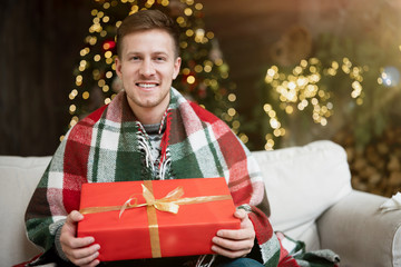 Fototapeta premium Young handsome man wearing warm sweater holding present sitting on the sofa in room decorated for celebrating the new year and christmas festive mood