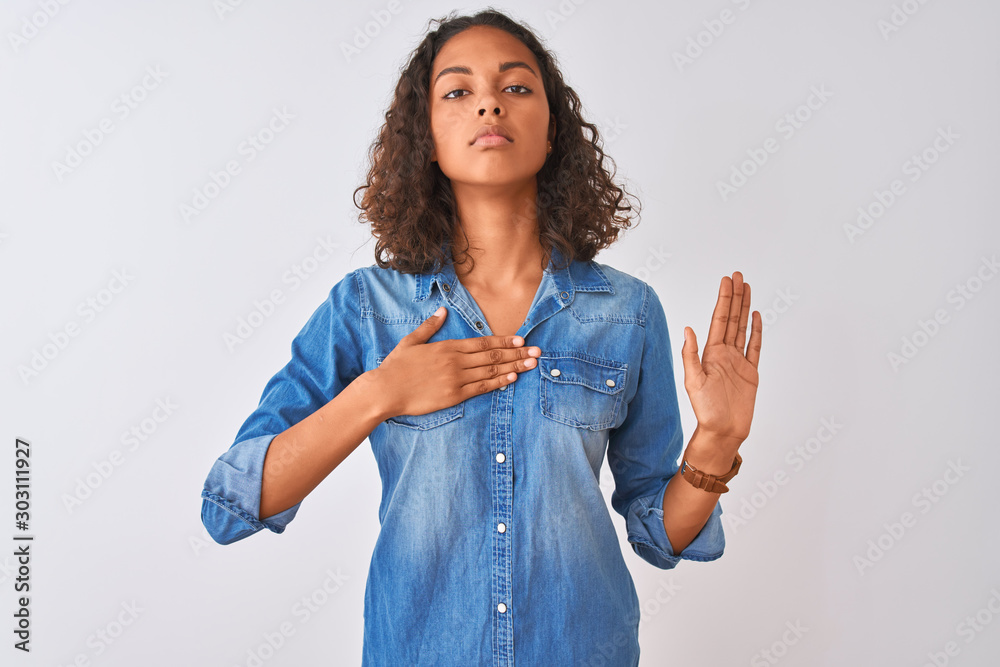 Wall mural Young brazilian woman wearing denim shirt standing over isolated white background Swearing with hand on chest and open palm, making a loyalty promise oath