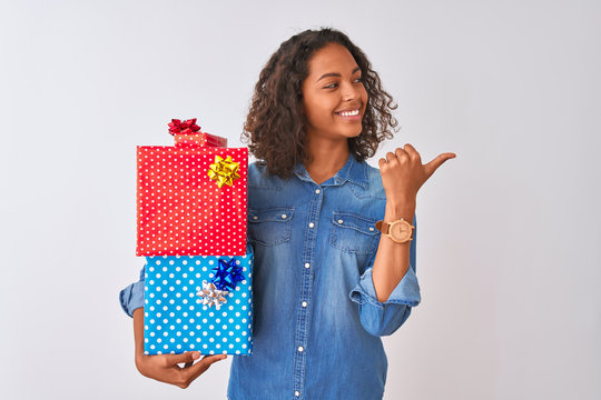 Young brazilian woman holding birthday gifts standing over isolated white background pointing and showing with thumb up to the side with happy face smiling