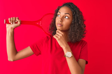 Naklejka premium Young brazilian sportswoman holding tennis racket standing over isolated red background serious face thinking about question, very confused idea