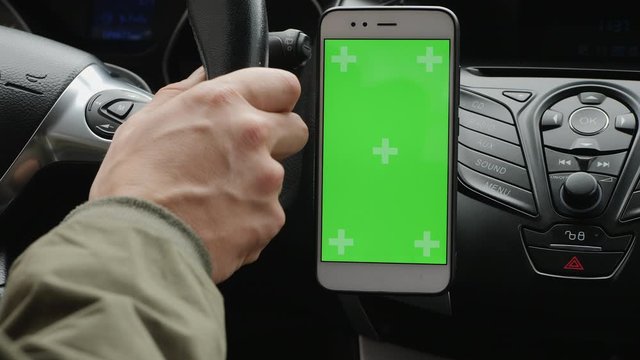Close-up hand of driver is driving a car while holding a steering wheel, uses a smartphone with a green screen is fixed on the dashboard. A man touches his finger to the screen center