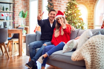 Young couple wearing santa claus hat sitting on the sofa around christmas tree at home smiling with happy face winking at the camera doing victory sign. Number two.