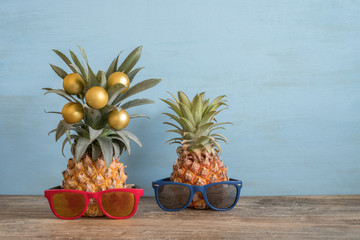 Pineapples in sunglasses with golden Christmas balls on wooden table. Free space for text.