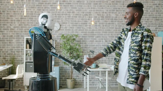 Side view of happy Afro-American businessman shaking hand to smart cyborg in office enjoying collaboration with artificial intelligence. Youth and technology concept.
