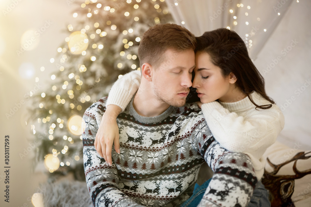 Wall mural lovely young couple beautiful woman and handsome man in cozy warm sweaters hugging on the sofa decorated for celebrating the new year christmas festive mood