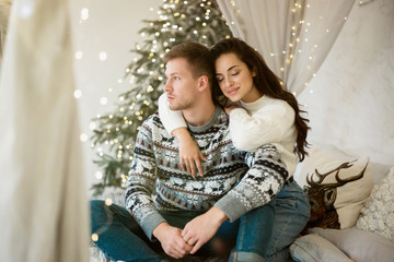 lovely couple beautiful wife and handsome husband both wearing warm sweaters cuddling hugging on the sofa decorated for celebrating the new year christmas festive mood