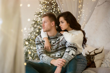 lovely young couple beautiful woman and handsome man both wearing cozy sweaters hugging on the sofa decorated for celebrating the new year christmas festive mood