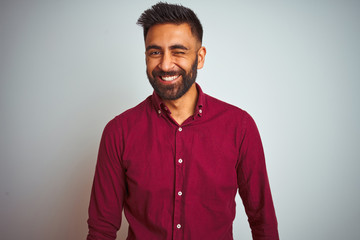 Young indian man wearing red elegant shirt standing over isolated grey background winking looking...