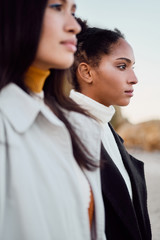 Close up two attractive girls in trench coats dreamily standing together by the sea